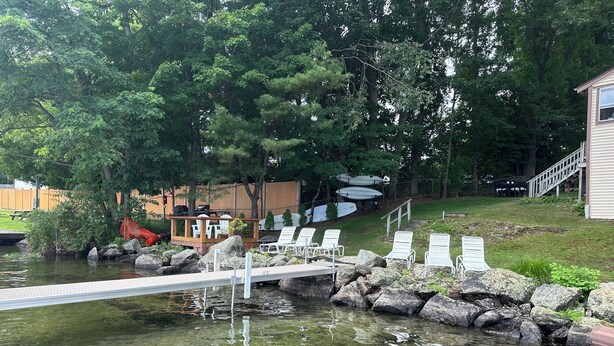 A clear pond with a wooden dock, surrounded by rocks and trees. There are white lounge chairs and a small wooden structure with umbrellas.