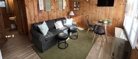 A cozy living room with a dark grey sofa, two black stools, a small round table, and a TV mounted on the wall.