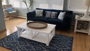 Una sala de estar con un sofá azul oscuro, una silla de mimbre, una mesa de centro blanca y una alfombra estampada.