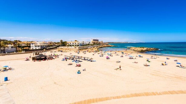 Una playa con una amplia zona arenosa, un litoral rocoso y un cielo azul despejado.