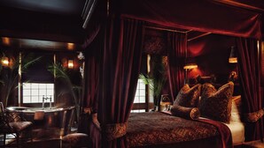Um quarto luxuoso com uma cama grande, almofadas elaboradas, um espaço de estar com uma poltrona e uma banheira.
