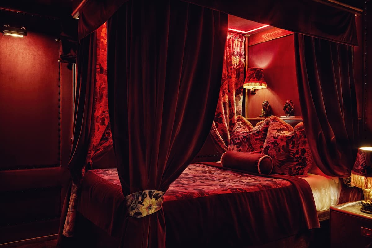 A room with a four-poster bed, red curtains, and a lamp on a bedside table.