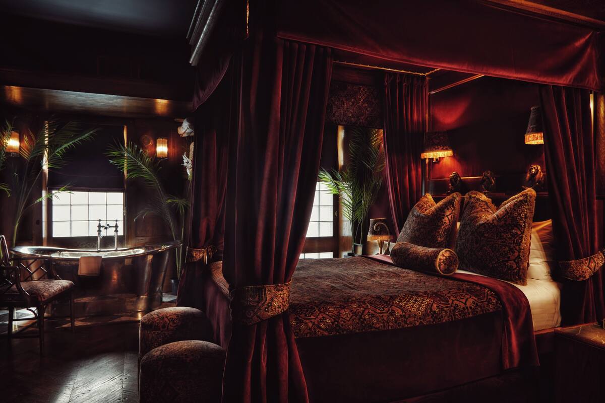 A luxurious bedroom with a large bed, ornate pillows, a sitting area with a chair, and a bathtub.