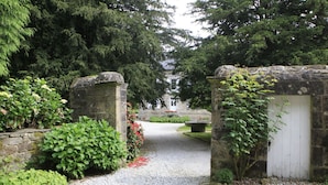 A gravel pathway leading to a stone gate with a white door, surrounded by lush greenery and a building in the background. - Le Manoir Du Poul Coeur de Bretagne B&B (Mellionnec)