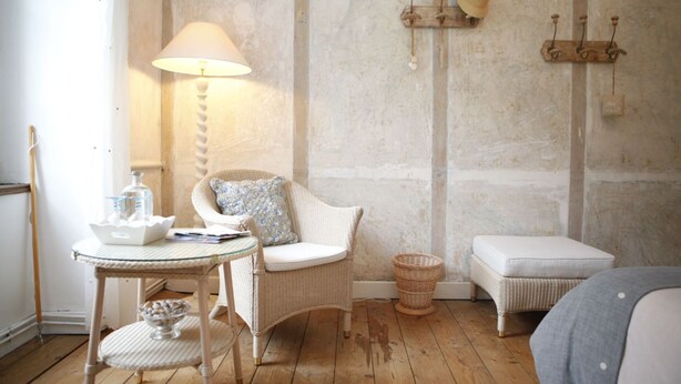 A cozy room with a wicker chair, a small round table with a lamp, and a white bench. - Le Manoir Du Poul Coeur de Bretagne B&B (Mellionnec)