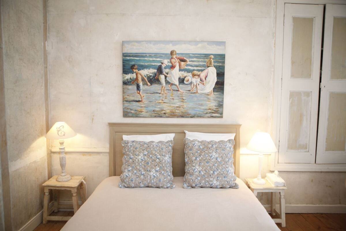 A bedroom with a large bed, two bedside tables, a nightstand, and a painting of a beach scene.