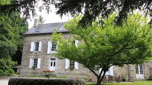 A stone building with white shutters, a large tree in front, and a stone planter with flowers. - Le Manoir Du Poul Coeur de Bretagne B&B (Mellionnec)