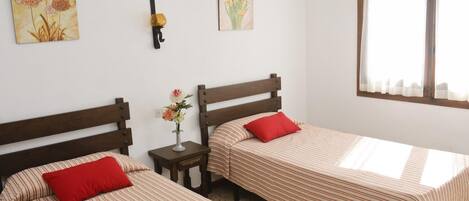 A room with two single beds, each with a red pillow and a striped bedspread. There is a small table with a vase of flowers between the beds. Two paintings are hung on the wall above the beds.