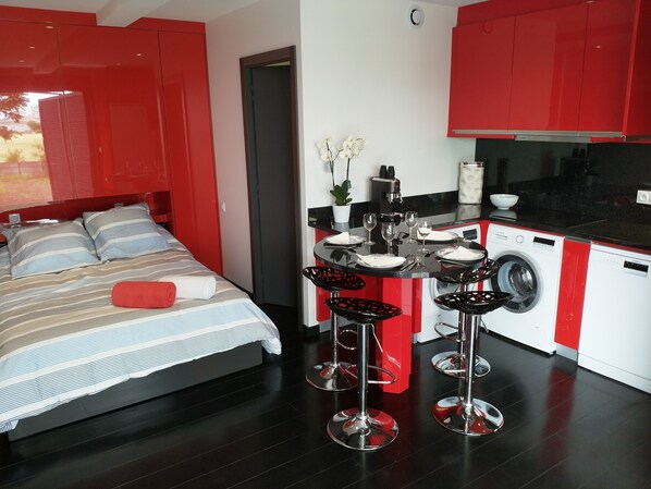 A modern hotel room with a bed, a small kitchen area, and a dining table with chairs.