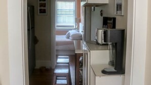 Un estrecho pasillo que conduce a un dormitorio con cama y ventana. Se ve un refrigerador a la izquierda y un microondas a la derecha.
