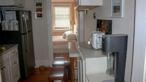 Una cocina estrecha con un refrigerador, una tostadora y un microondas. Hay un banco en el centro y una ventana con persianas en el fondo.