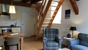 Un salon chaleureux avec un fauteuil à carreaux, un canapé bleu et une table basse en bois. On aperçoit une cuisine avec une cuisinière et des placards au fond.