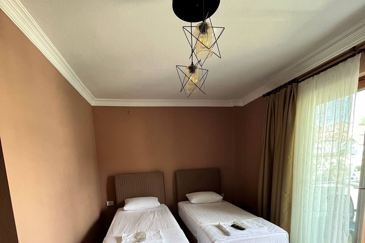 Ein Hotelzimmer mit zwei Einzelbetten, Holzboden, einer Deckenleuchte und einem Fenster mit Vorhängen.