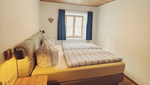 Ein kleiner, gemütlicher Raum mit Holzbalkendecke, einem Einzelbett mit gemustertem Überwurf, einem Nachttisch und einem Fenster mit Vorhängen.