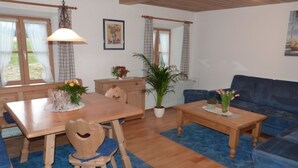 Ein gemütliches Wohnzimmer mit Esstisch aus Holz, blauem Sofa und blauem Teppich. Auf dem Tisch stehen Blumentöpfe und ein Stuhl mit herzförmiger Aussparung.