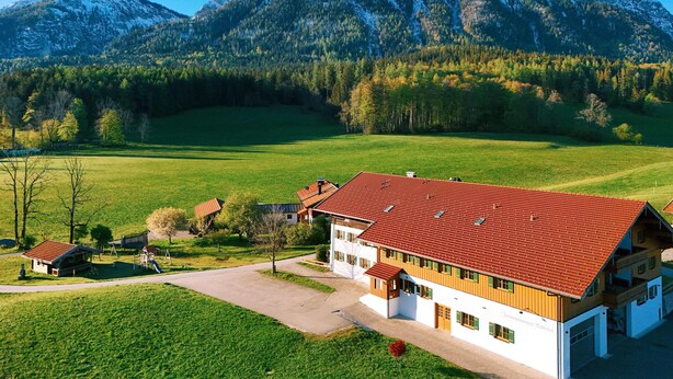 Ein zweistöckiges Gebäude mit rotem Ziegeldach, umgeben von grünen Feldern und einer Bergkette im Hintergrund.