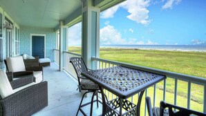 A covered porch with wicker furniture and a table, overlooking a grassy field and the ocean. - Salty Air Retreat - Fabulous Beach Views - Brand New Elevator (Galveston)