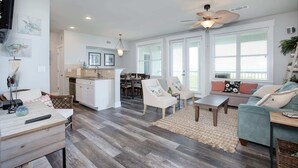 A spacious living area with a wooden floor, a ceiling fan, and a dining area with a kitchen in the background. - Salty Air Retreat - Fabulous Beach Views - Brand New Elevator (Galveston)