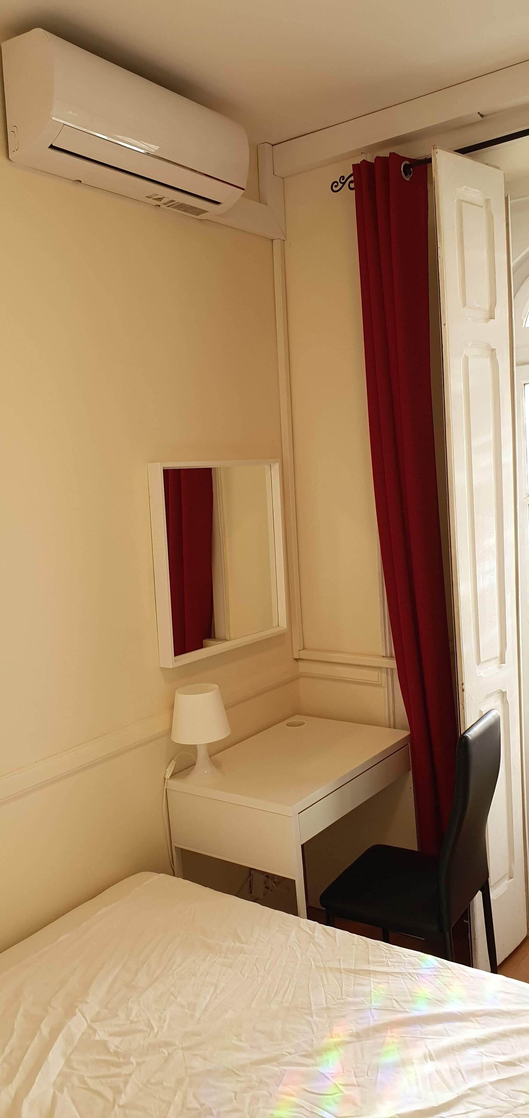 A hotel room with a bed, a desk, a chair, a mirror, an air conditioning unit, and a red curtain.