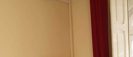 A hotel room with a bed, a desk, a chair, a mirror, an air conditioning unit, and a red curtain.