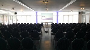 Una sala conferenze con file di sedie rivolte verso uno schermo per presentazioni, ampie finestre e luci a soffitto.