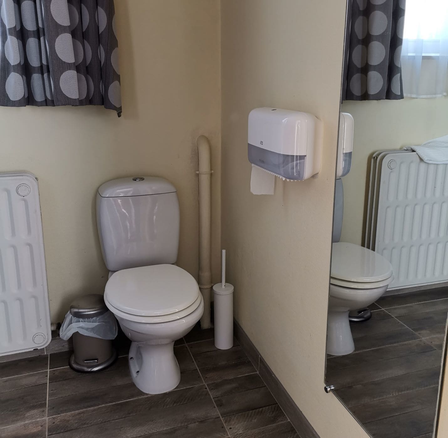 A bathroom with two toilets, a hand dryer, a trash bin, and a radiator.
