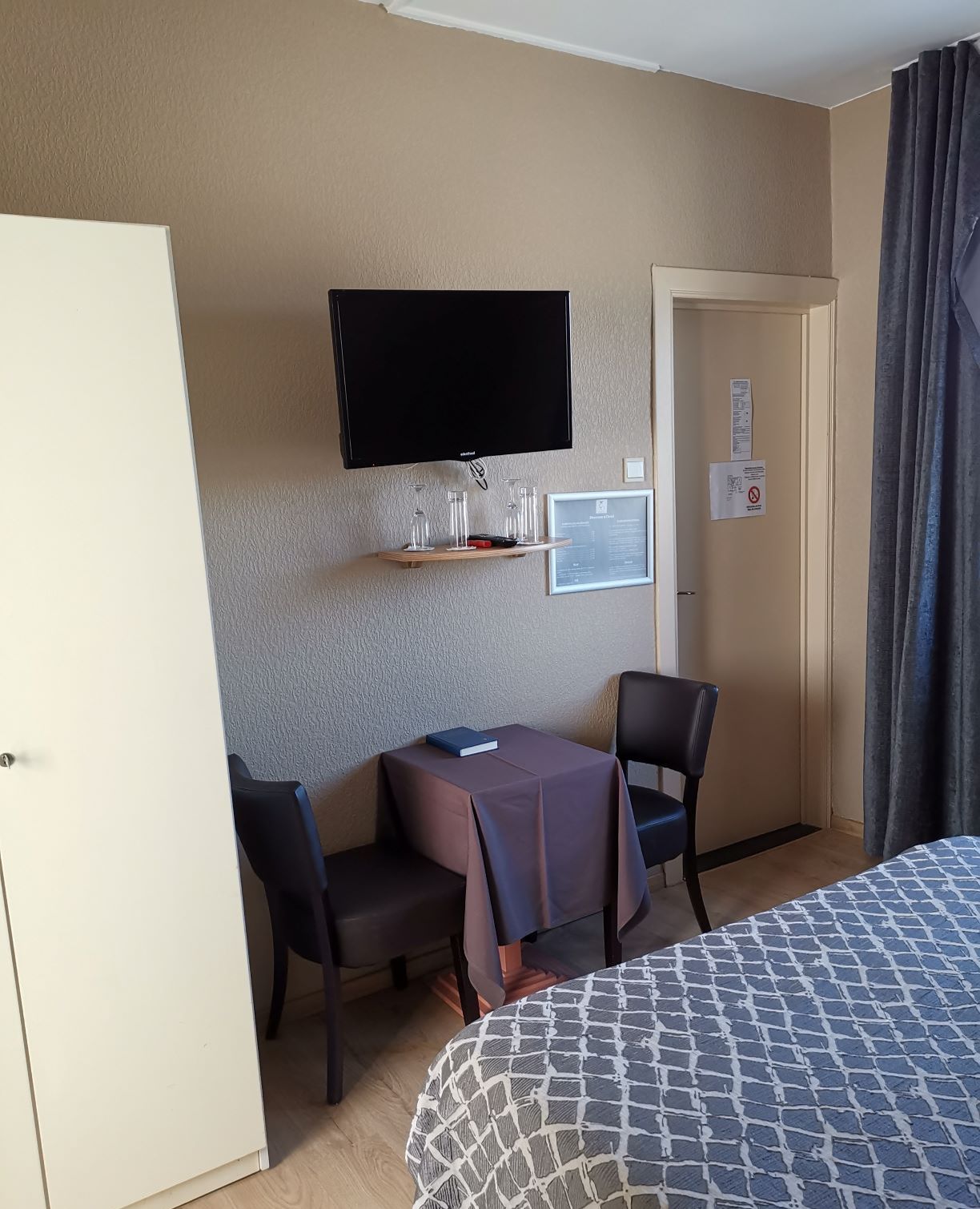A hotel room with a bed, a desk with a chair, a television mounted on the wall, and a door.