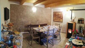 Un'area pranzo con un tavolo in legno apparecchiato per un pasto, circondato da sedie. Un televisore è montato a parete e sullo sfondo si nota un camino.