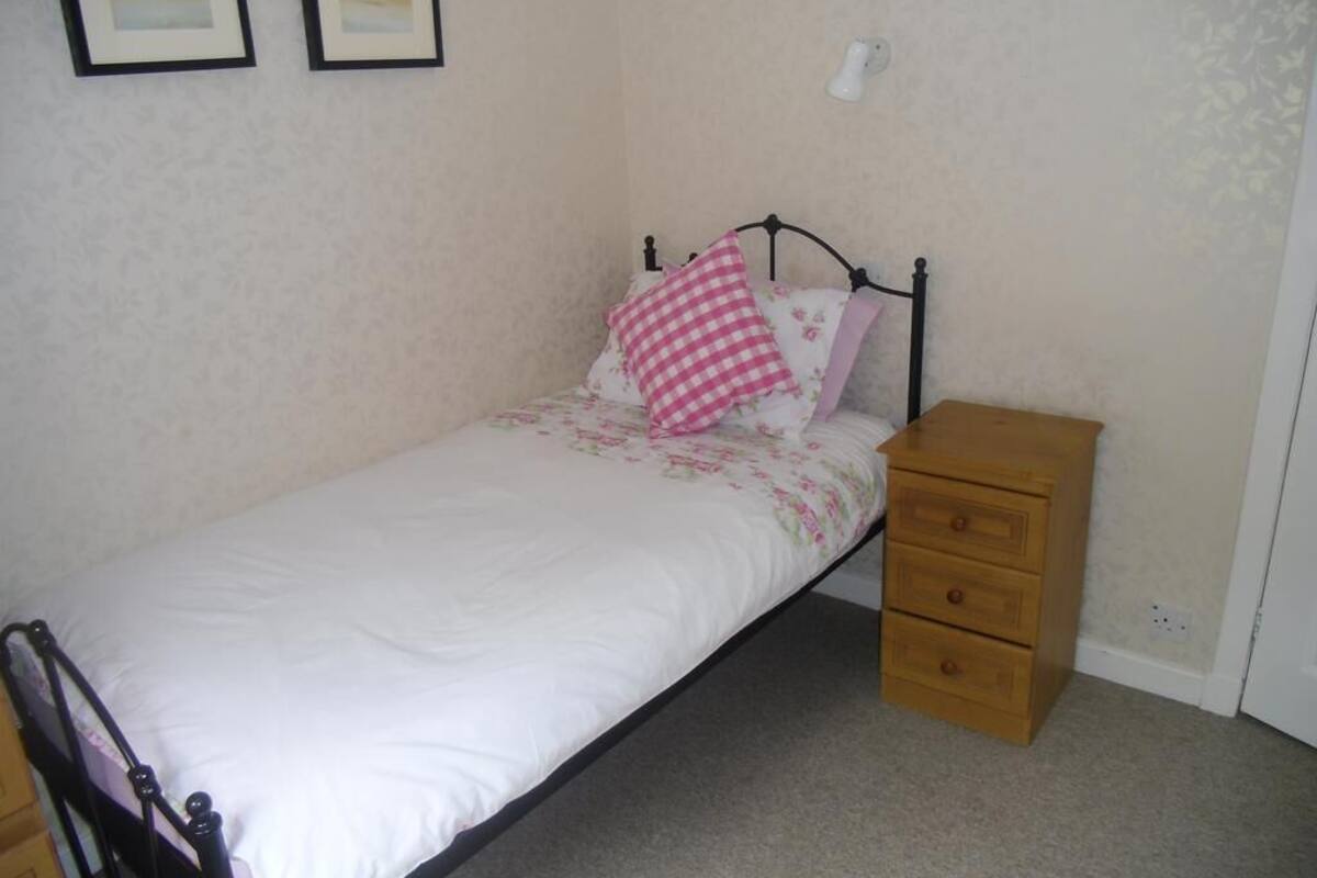 A single bed with a white mattress and floral bedding, a wooden nightstand with drawers, and a wall-mounted light fixture.