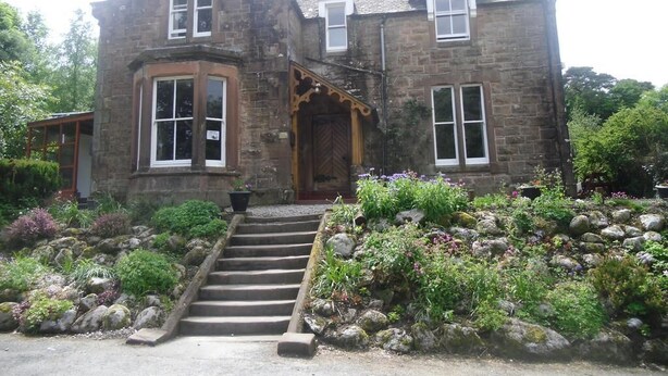 A stone building with a steep staircase leading to a wooden door, surrounded by a garden with various plants and rocks. - The Ormidale Hotel (Isle of Arran)