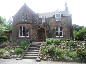 A stone building with a steep staircase leading to a wooden door, surrounded by a garden with various plants and rocks. - The Ormidale Hotel (Isle of Arran)