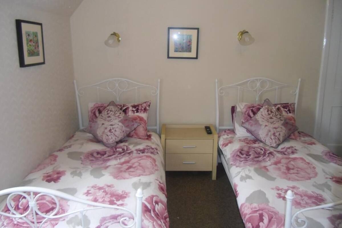 A bedroom with two single beds, each with a floral bedspread and matching pillows. There is a small bedside table with a lamp and a remote control. The room has a sloped ceiling and two framed pictures on the wall.