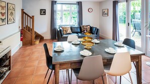 A dining area with a table set for four, a sofa with decorative pillows, a staircase, and a view through large windows. - Ship Bungalow (Minehead)