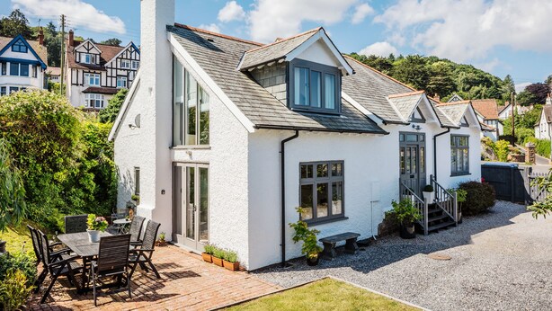 A white house with a sloped roof, a chimney, and a small balcony. There is a paved patio with a table and chairs. - Ship Bungalow (Minehead)