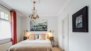 A bedroom with a bed, two bedside tables with lamps, a chandelier, a painting, and a door. - Ship Bungalow (Minehead)