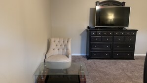 Una habitación con una mesa de vidrio, un sillón tapizado, un aparador negro y un televisor encima.