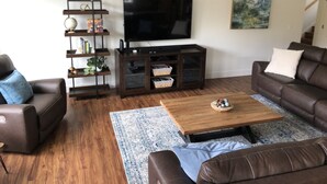 A living room with a brown leather sofa, a coffee table, a TV, a bookshelf, and a painting on the wall. - CHARMING AND SPECTACULAR RIVERFRONT HOUSE, BEAUTIFUL IN ANY SEASON OF THE YEAR! (Leaburg)