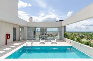 A rooftop swimming pool with clear blue water, surrounded by white walls and a glass door leading to a balcony with a view of the city. - OXO Bertoni Luxury BBQ & Pool (Asunción)