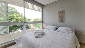 A modern bedroom with a large window, a bed with white bedding, and a wall-mounted macrame piece. - OXO Bertoni Luxury BBQ & Pool (Asunción)