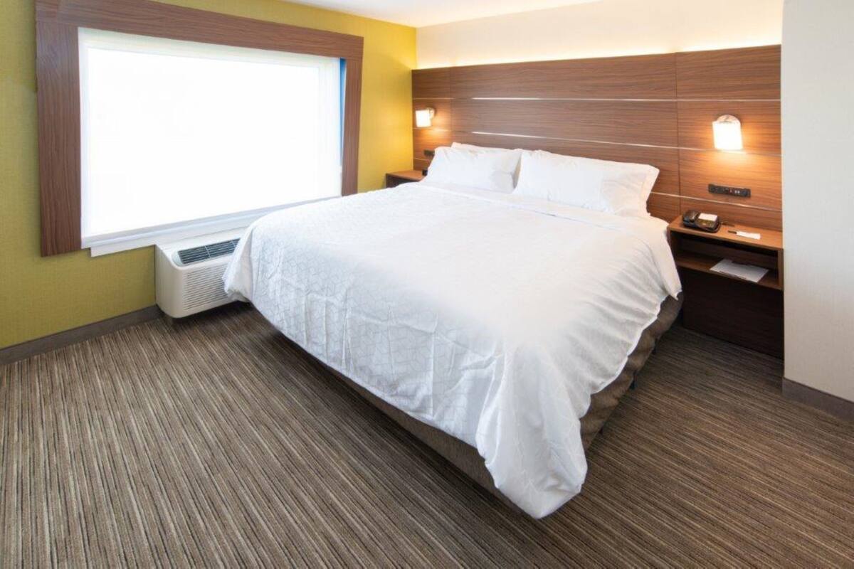 A hotel room with a large bed, a window with blinds, a wall-mounted air conditioner, a bedside table with a lamp, and a nightstand with a phone and a notepad.