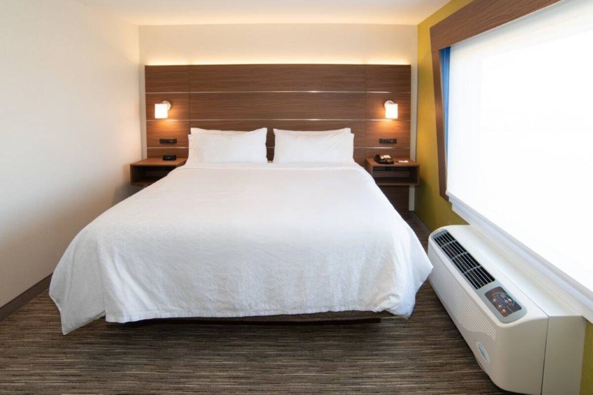 A hotel room with a large bed, two bedside tables with lamps, a wall-mounted air conditioner, and a window with curtains.
