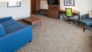 A hotel room with a blue sofa, a wooden desk with a television, a chair, and a small table. - Holiday Inn Express & Suites North Battleford by IHG (North Battleford)