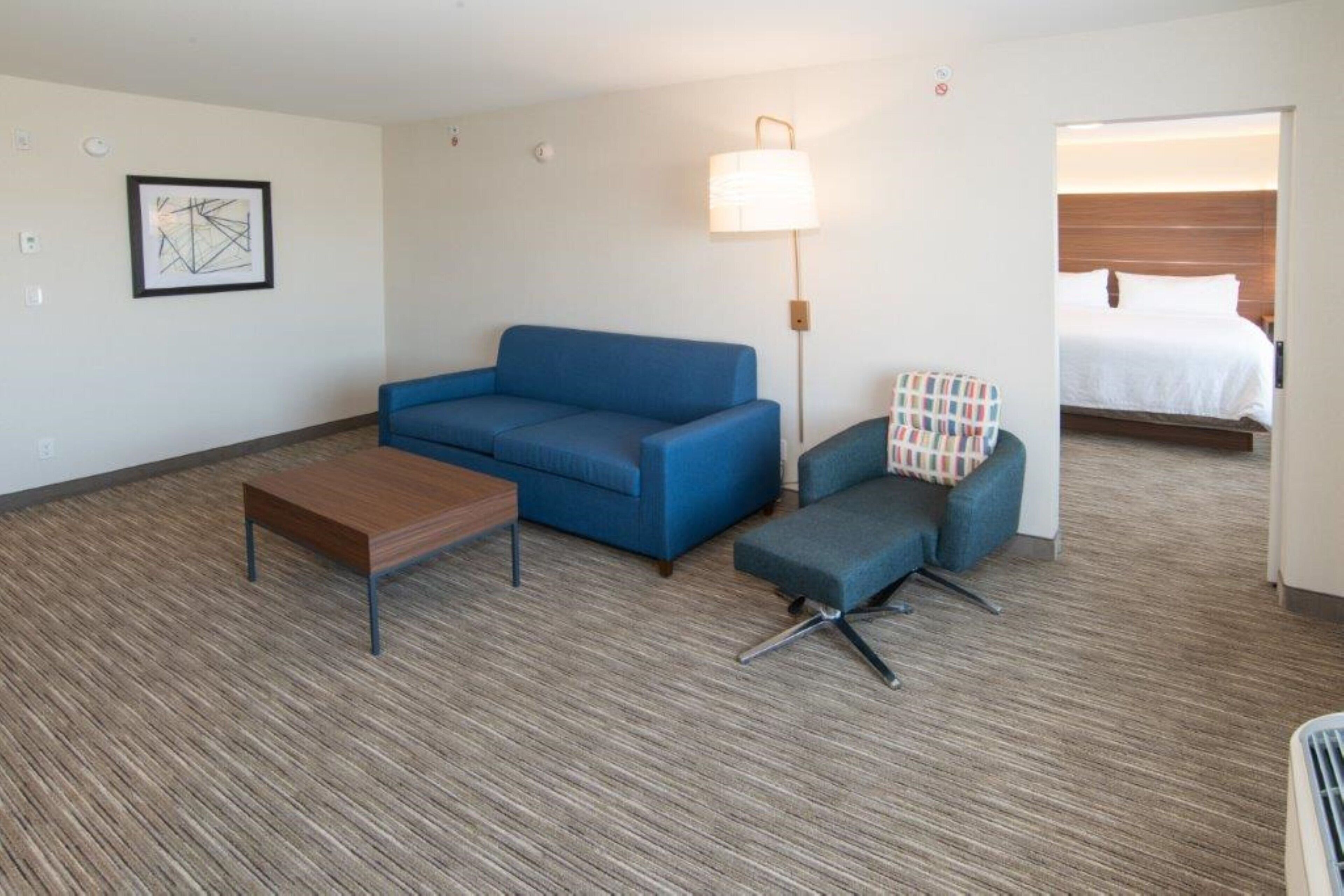 A hotel room with a blue sofa, a wooden table, a patterned armchair, and a bed with white linens.
