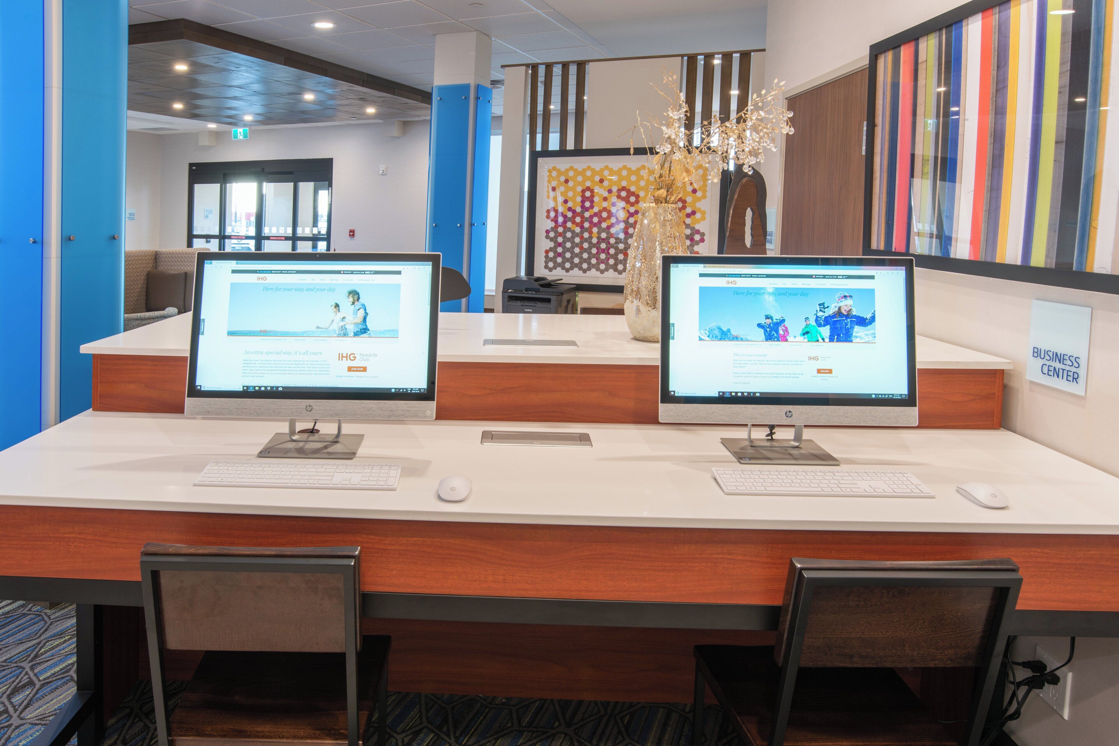 Un mostrador de recepción con dos monitores HP mostrando el sitio web del Club de Recompensas IHG, un letrero que indica centro de negocios y un jarrón decorativo con ramas.