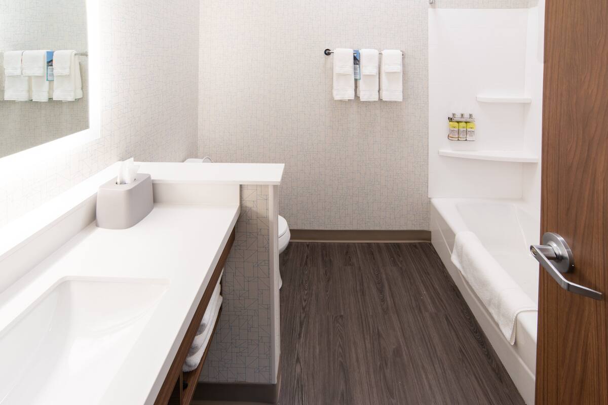 A modern bathroom with a white sink, toilet, and bathtub. There are towels hanging on the wall and a shelf with toiletries.