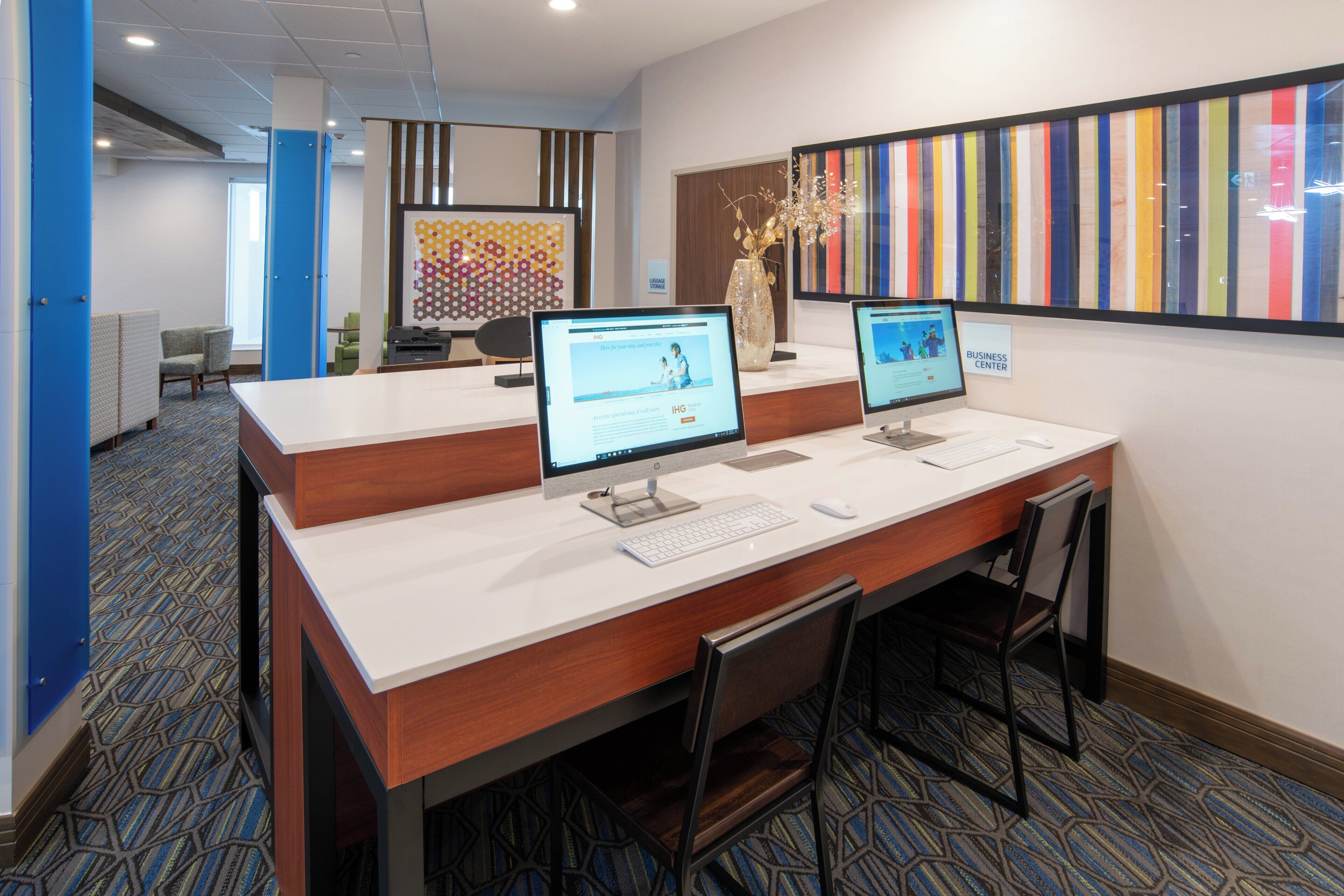 Una recepción con dos monitores de computadora, un florero con flores y una obra de arte con motivos coloridos a rayas.