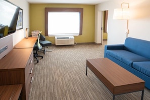 Une chambre d’hôtel avec un bureau en bois, un canapé bleu, une télévision, un appareil de climatisation et une fenêtre avec des stores.