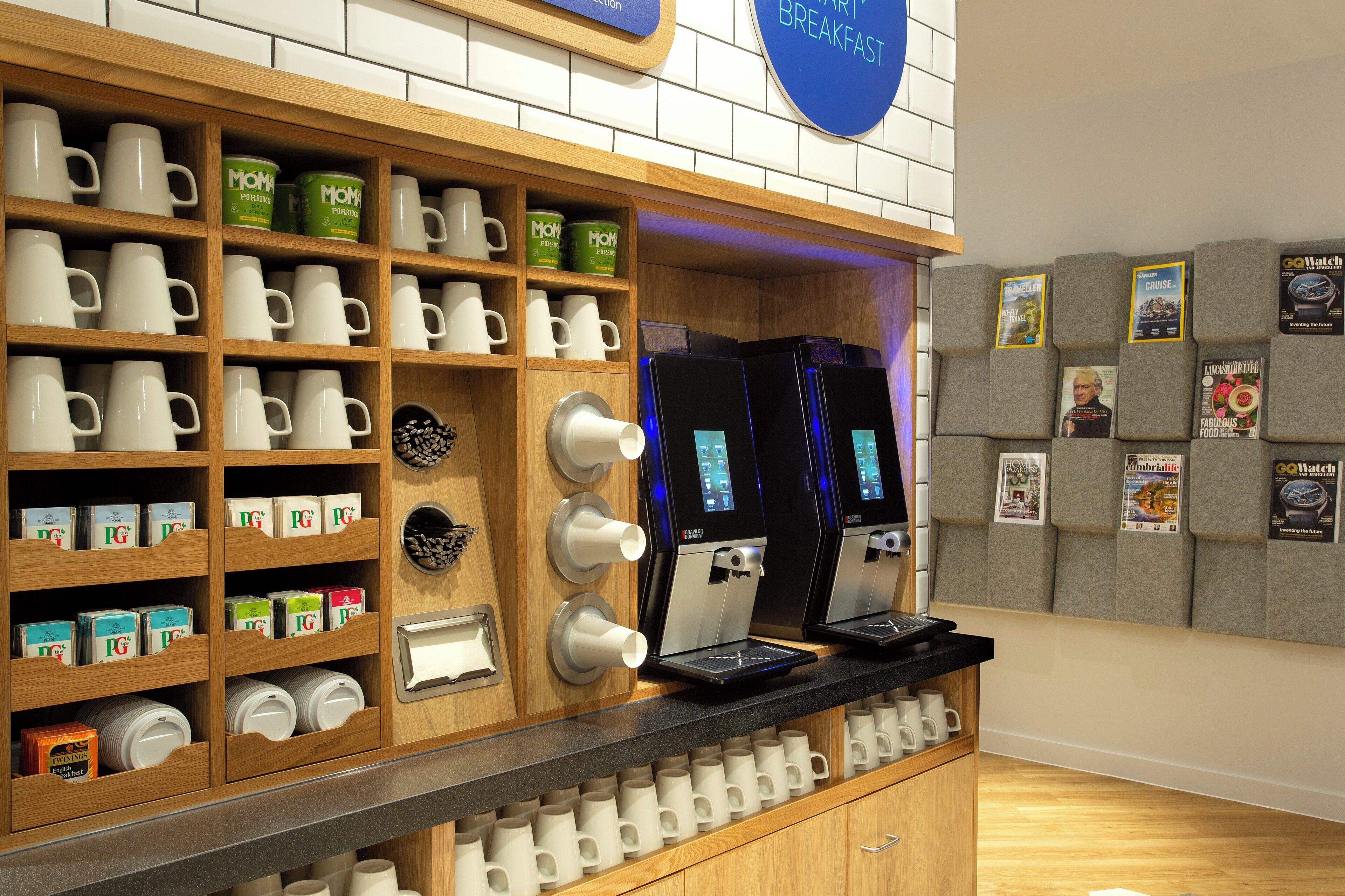 A coffee station with multiple coffee machines, a variety of coffee cups, and a display of magazines.