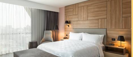 A modern hotel room with a large bed, a bedside table with a lamp, a chair, and a view of the city through large windows.