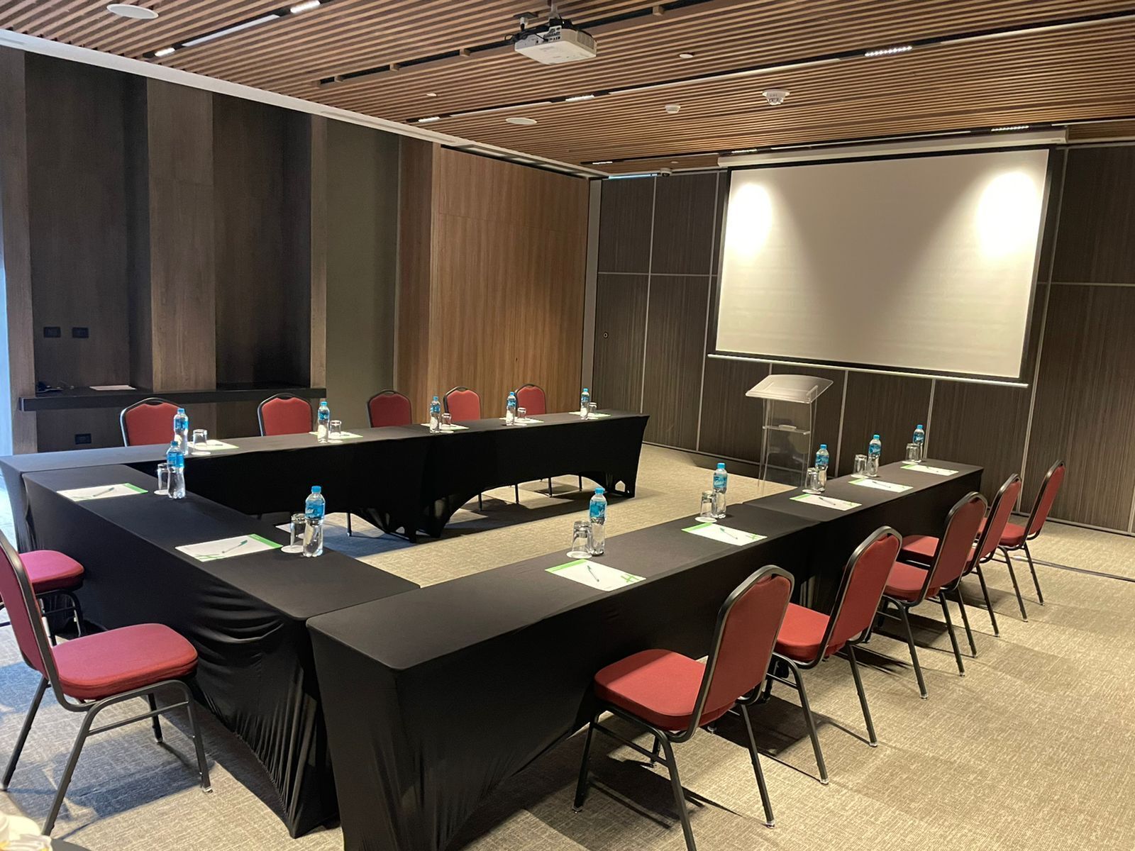 A conference room with a long table, red chairs, water bottles, and a projector screen.